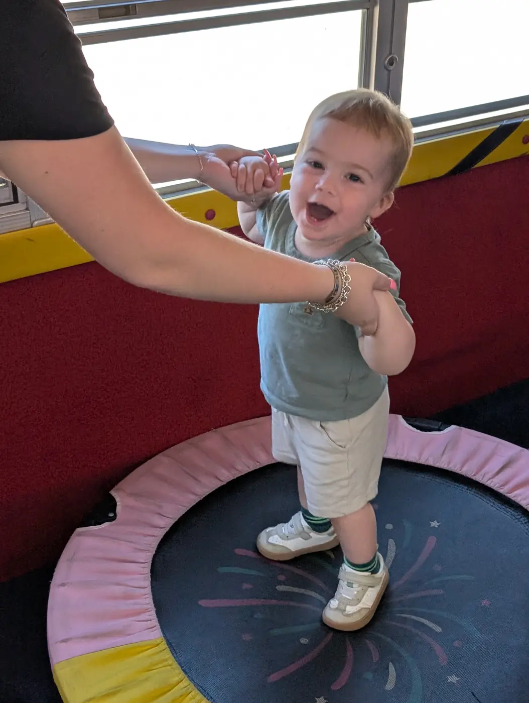 A joyful toddler bouncing on a trampoline with a teacher at Blossom Station in Acton, MA